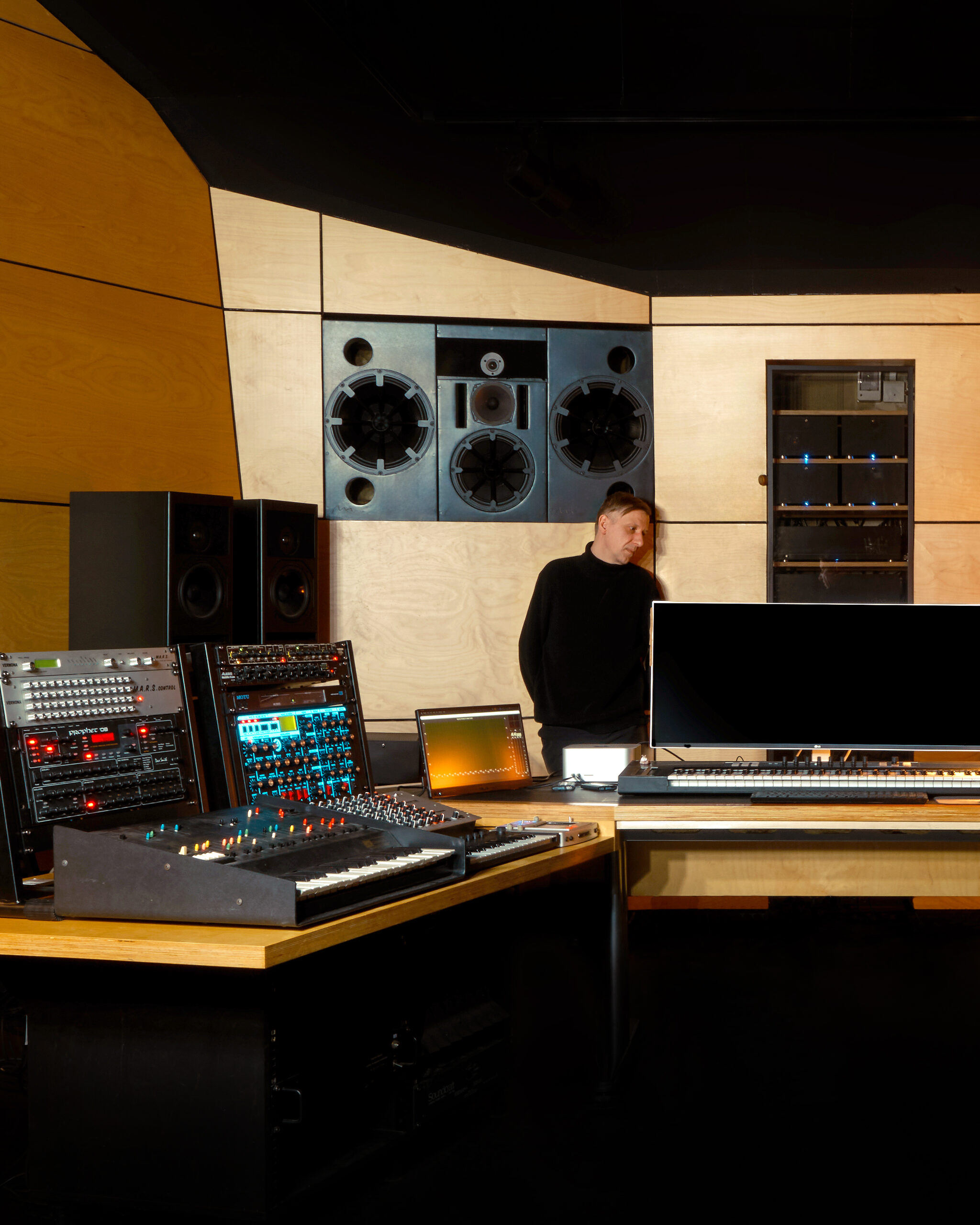 Ten Walls standing in a wood-paneled music studio among synthesizers, studio monitors and production equipment